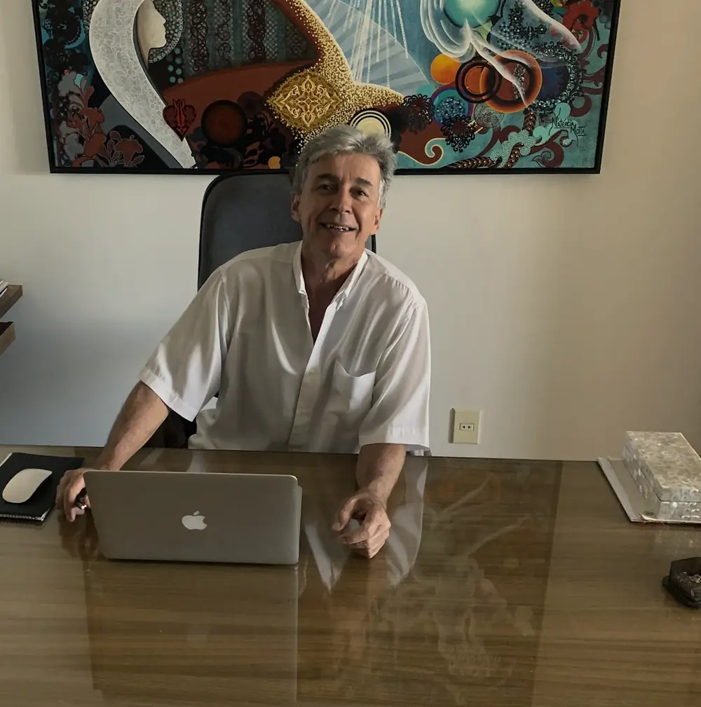 Luiz Fernando sorrindo à mesa do consultório, com o notebook aberto e um quadro colorido atrás